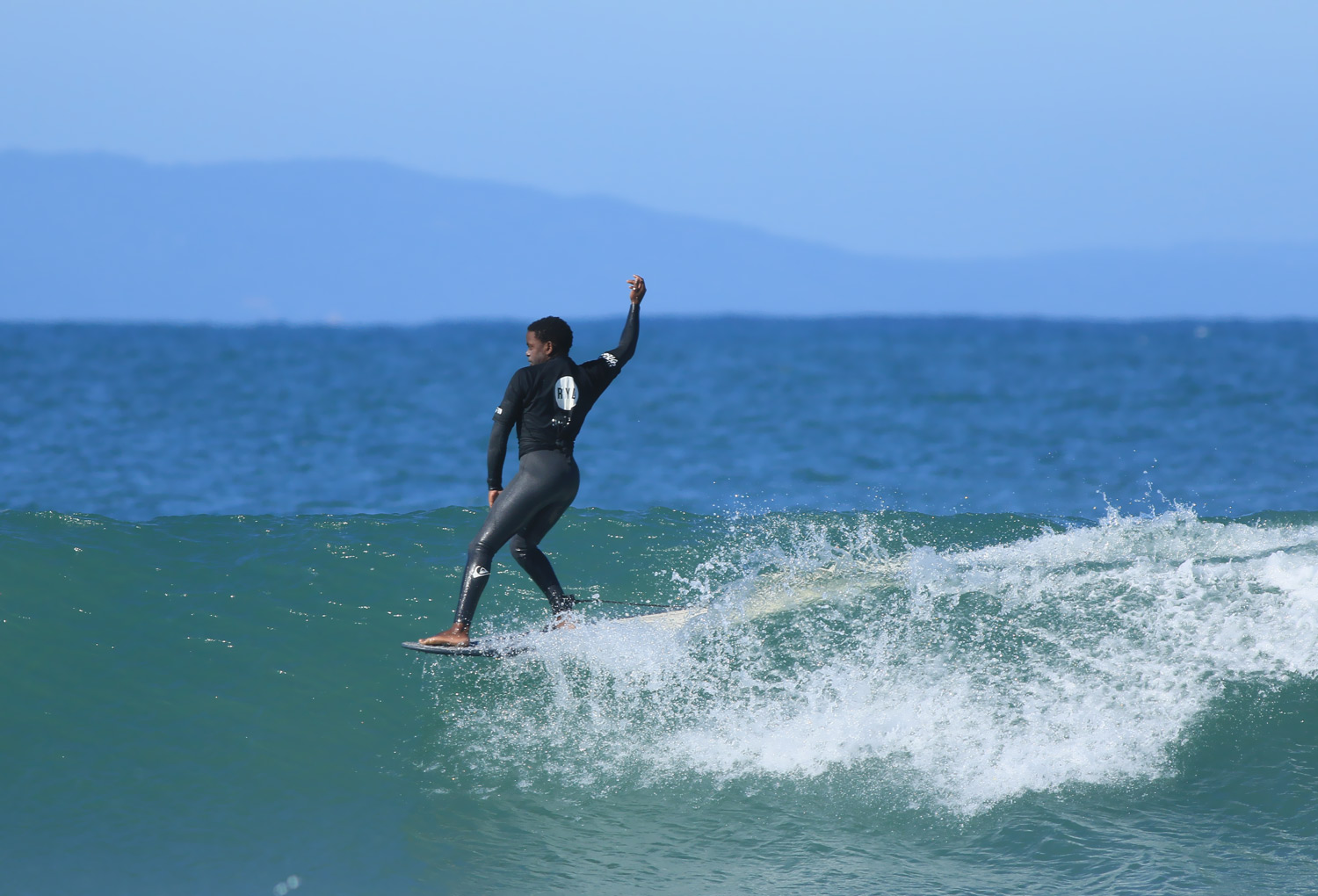 A young surfer riding a wave during a competition in South Africa, performing a hang five, wearing a black wetsuit.
DE: Ein junger Surfer in einem schwarzen Neoprenanzug reitet während eines Wettbewerbs in Südafrika auf einer Welle und vollführt einen Hang Five.
NL: Een jonge surfer rijdt op een golf tijdens een wedstrijd in Zuid-Afrika, terwijl hij een hang five uitvoert in een zwart wetsuit.
