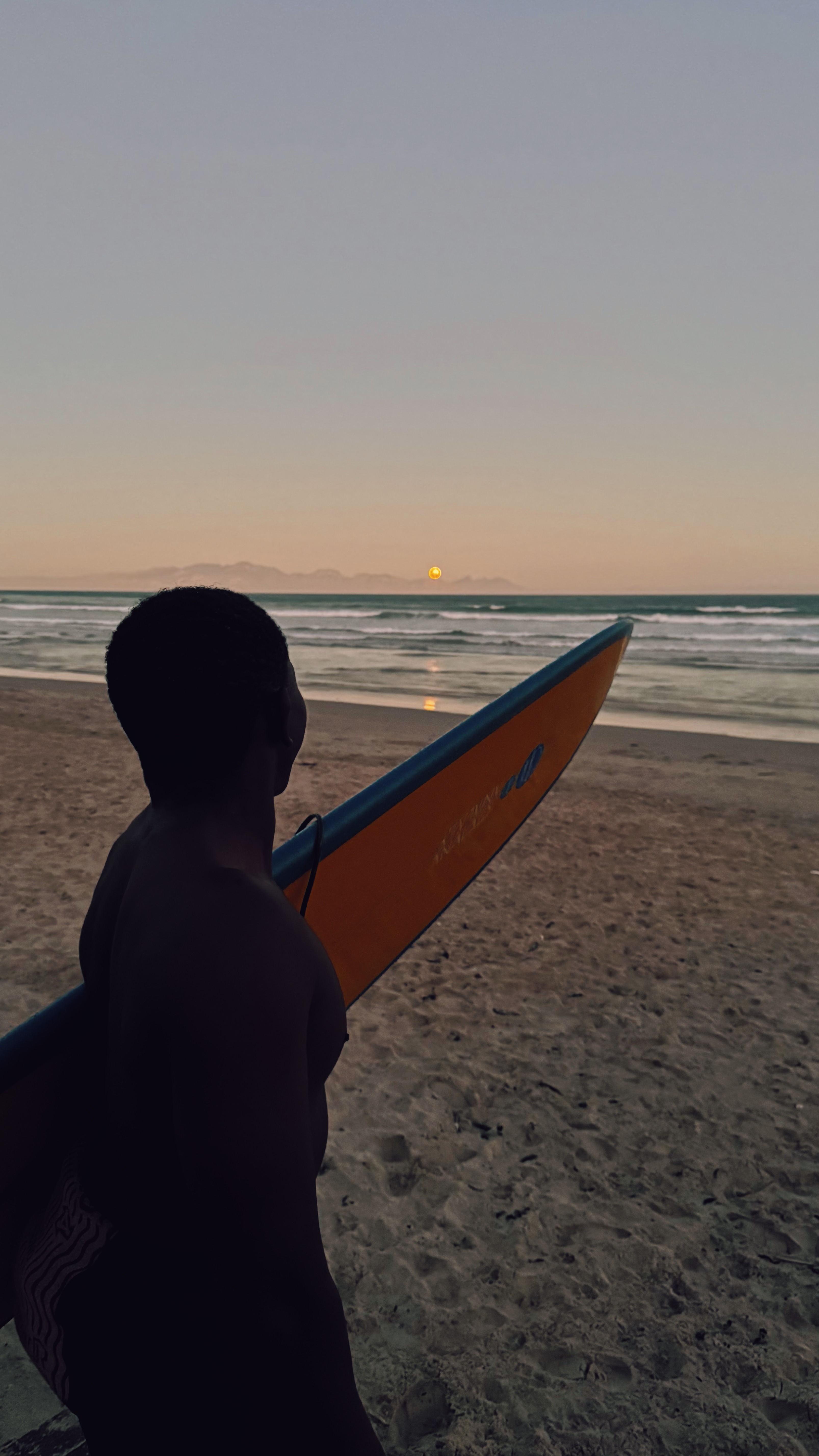 A surfer holding a surfboard while watching the sunset over the ocean on Muizenberg beach in Cape Town
DE: Ein Surfer hält ein Surfbrett, während er den Sonnenuntergang über dem Meer am Strand von Muizenberg in Kapstadt beobachtet
NL: Een surfer houdt een surfplank vast terwijl hij kijkt naar de zonsondergang over de oceaan op het strand van Muizenberg in Kaapstad