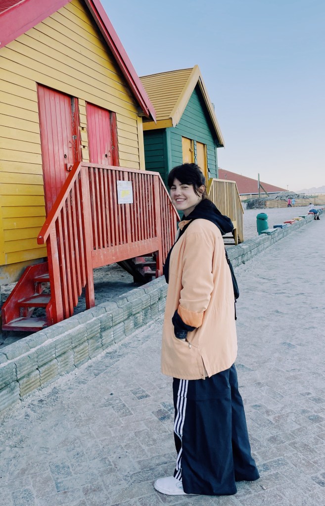 A female person smiling and standing on a pathway near legendery colored beach huts in Muizenberg, Cape Town.
