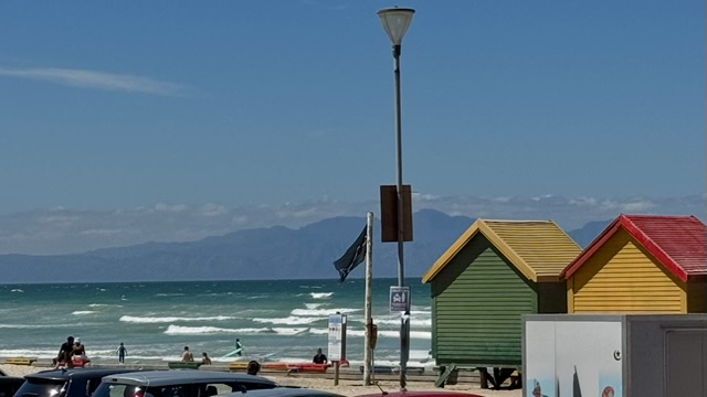 A view of Muizenberg beach featuring colorful beach huts, waves breaking in the ocean, and a black flag indicating caution against potential dangers.