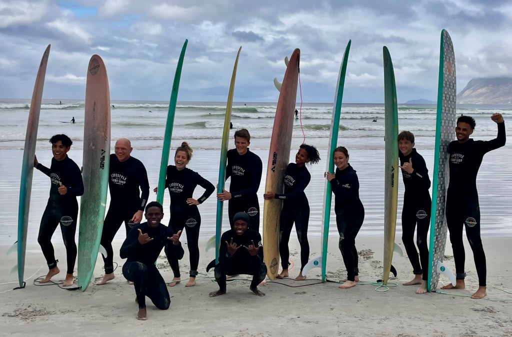 A group of beginner surfers posing and smiling at Muizenberg beach, Cape Town, wearing black wetsuits and holding colorful surfboards, with waves and clouds in the background. The 2 surf coaches are posing in front of the group, smiling.
