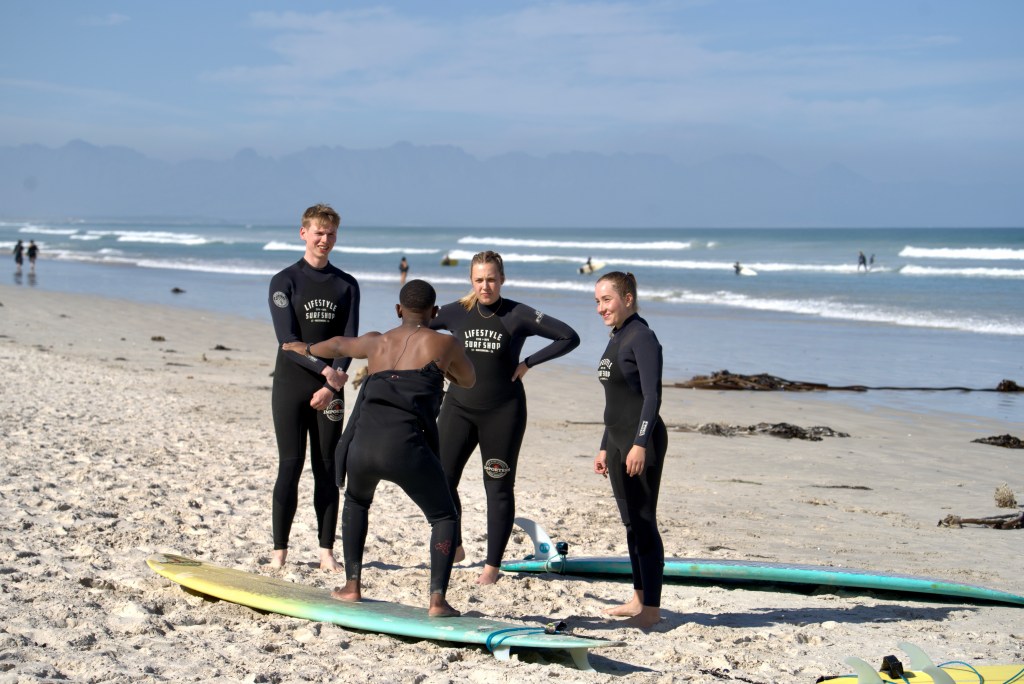 A surf coach standing on a soft-top surfboard at Muizenberg Beach, Cape Town, explaining the pop up to a group of beginner surfers.