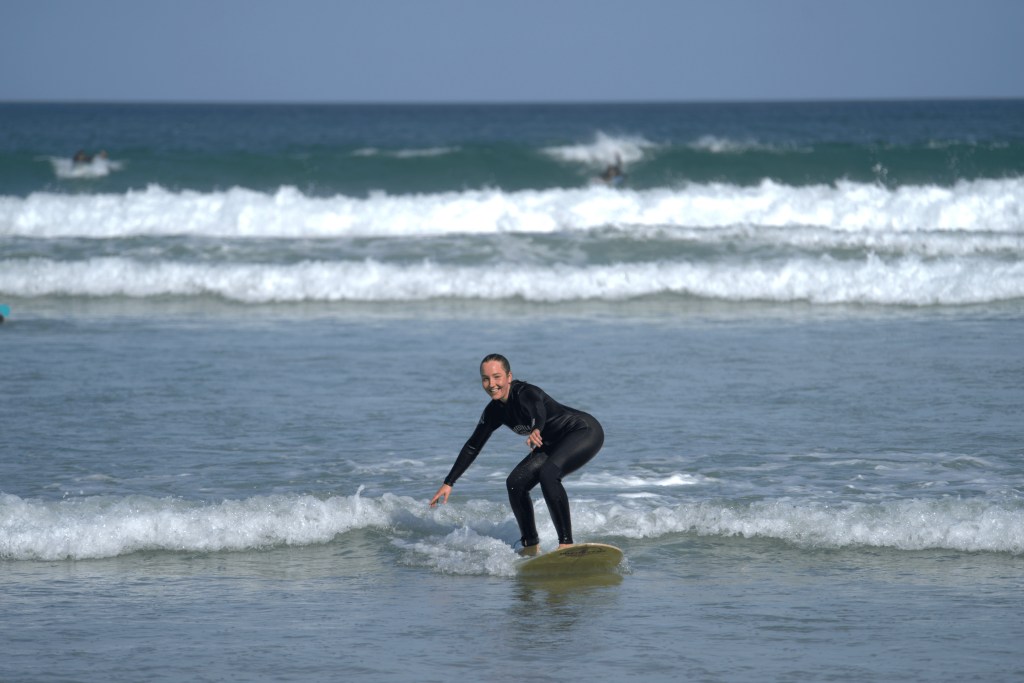 A smiling beginner surfer in a black wetsuit rides a small wave on sunny Muizenberg Beach in Cape Town.