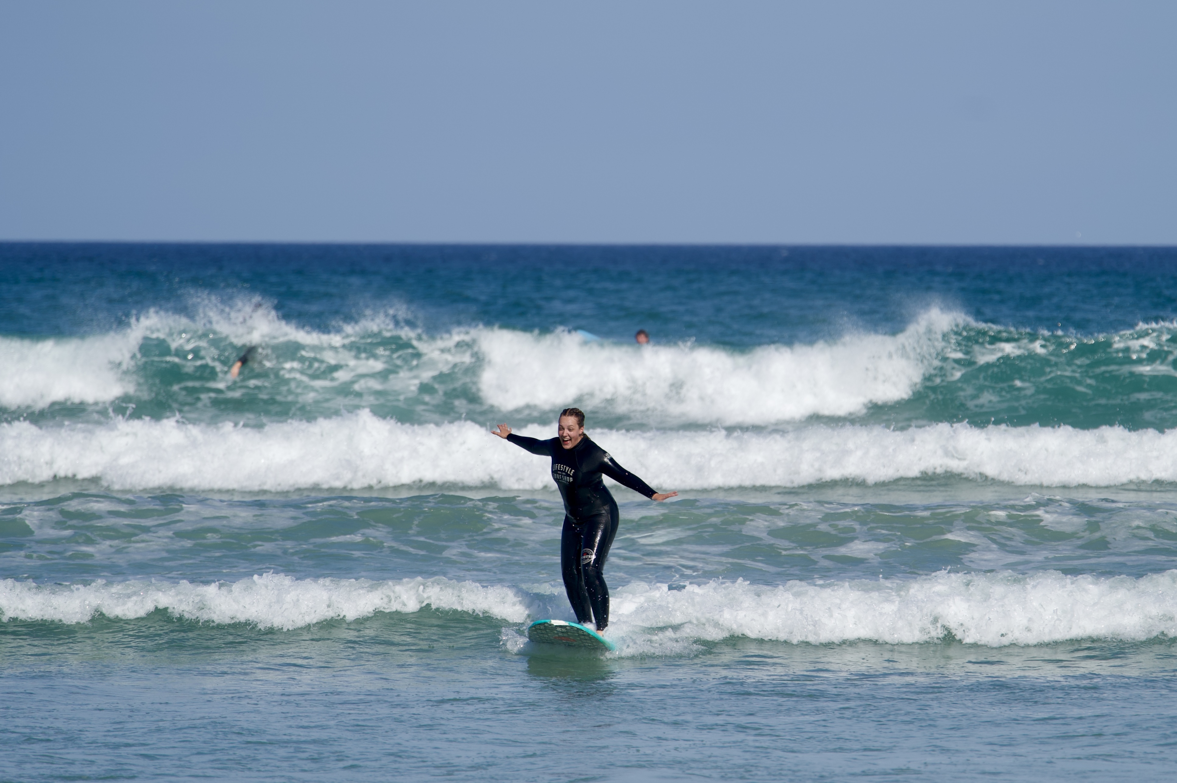 A beginner surfer surfing for the first time, standing on the soft-top surfboard, happy and excited. Beautiful ocean waves of Muizenberg, Cape Town, in the background, blue sky.
