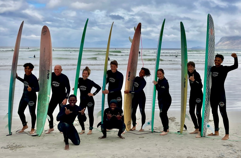 Group of beginner surfers posing on Muizenberg beach in Cape Town with surfboards and their surf coaches in front, wearing matching wetsuits, smiling and having fun.