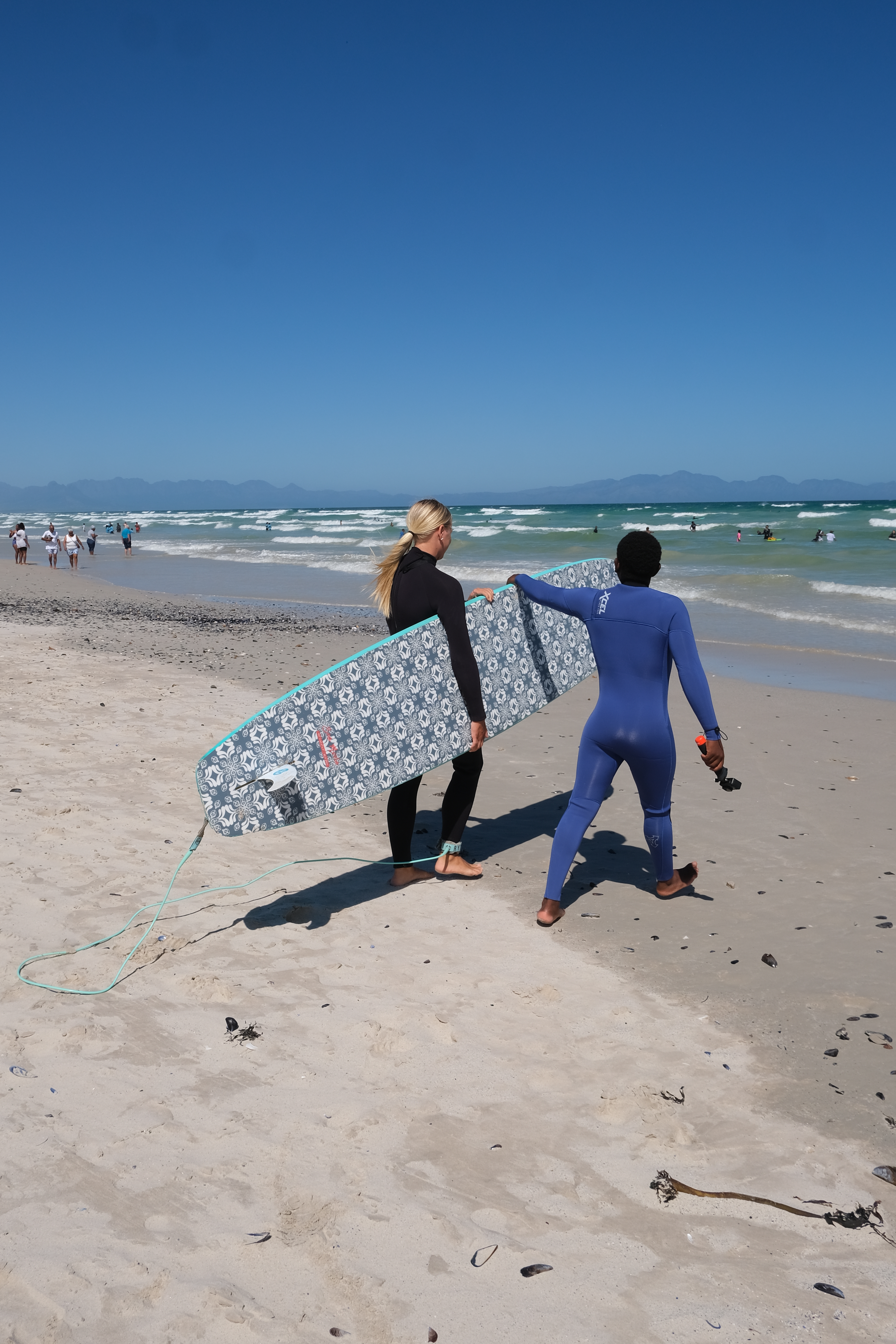 A female beginner surfer carrying a softtop surfboard, her surf coach helps her carry it. They are heading towards the water to start the surf lesson. Muizenberg Beach, Cape Town, South Africa