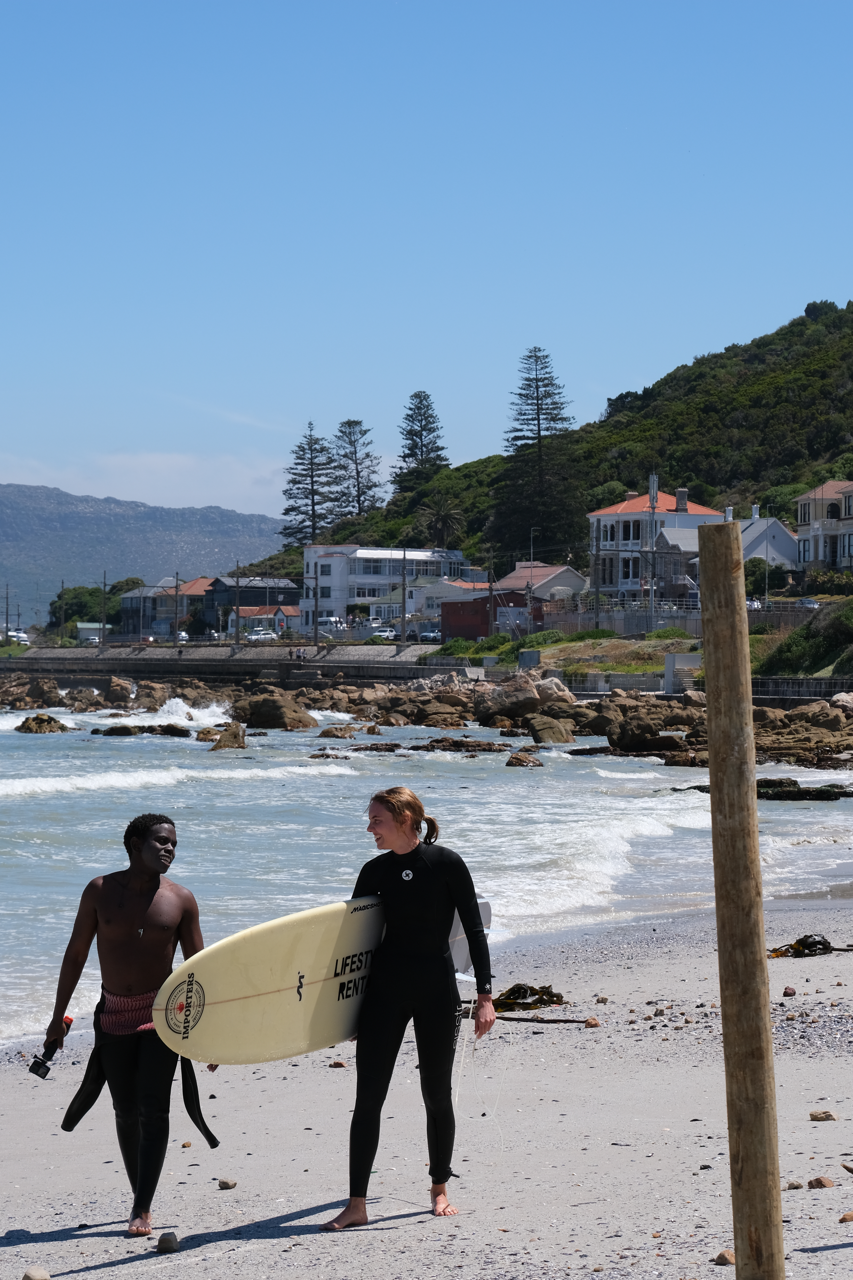 A beginner surfer carrying a surfboard, talking happily to her surf coach. They just got out of the water after the beginner lesson at Muizenberg Beach, Cape TOwn, South Africa